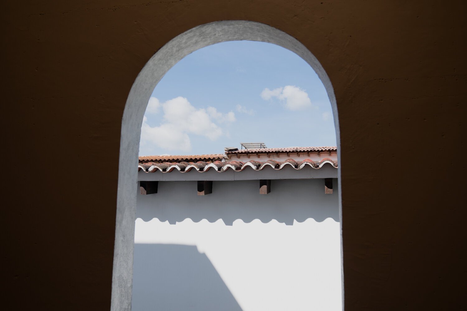 Vue d'un toit en tuiles à travers une arche, ciel bleu et nuages en arrière-plan.