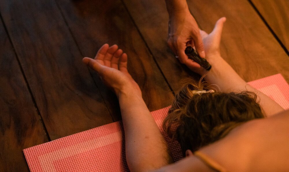 Personne allongée sur le ventre sur un tapis de yoga rose, bras étendus, tandis qu'une autre personne applique une petite pierre noire sur le bras.