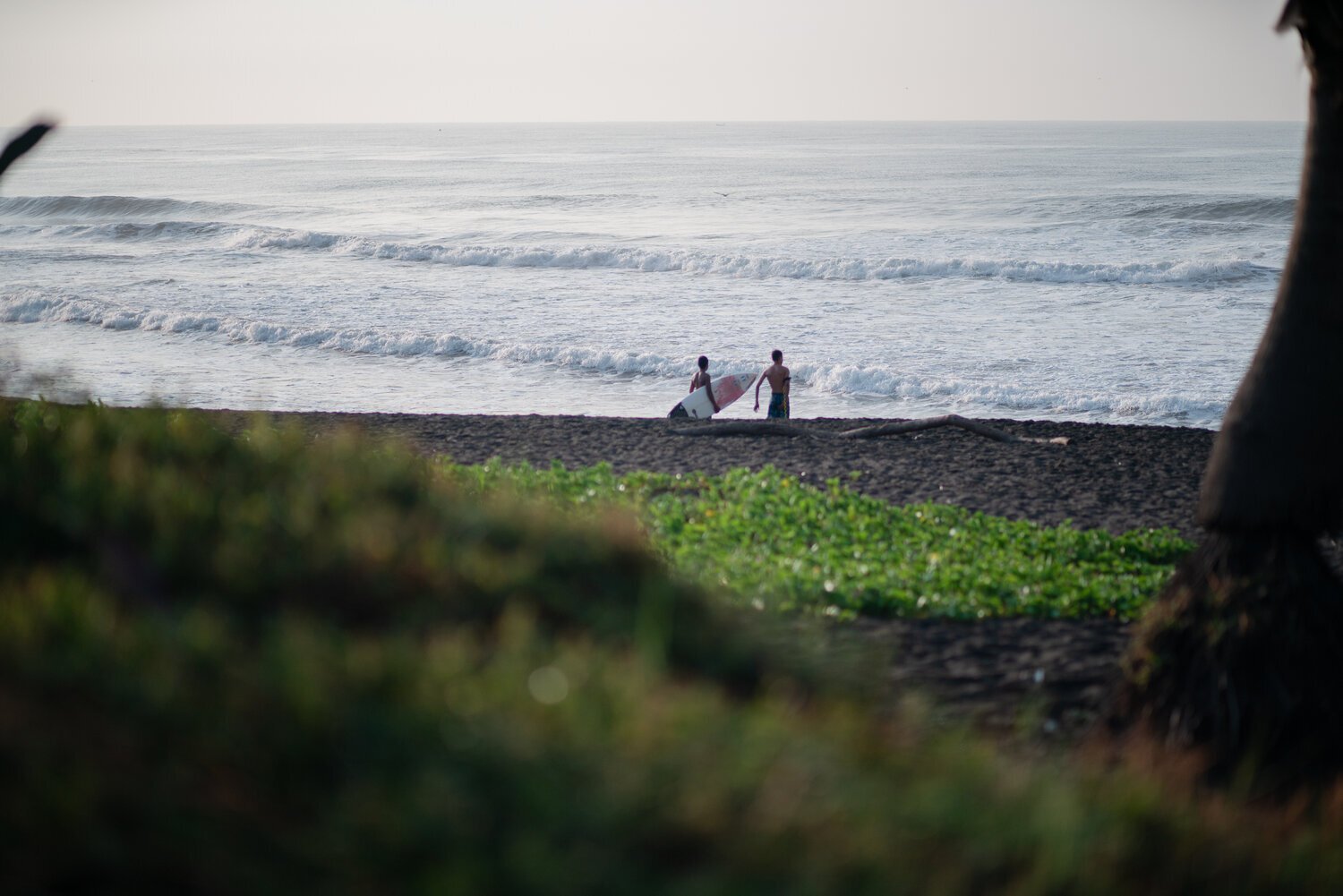 Deux personnes avec des planches de surf sur une plage face à l'océan, avec des vagues visibles.