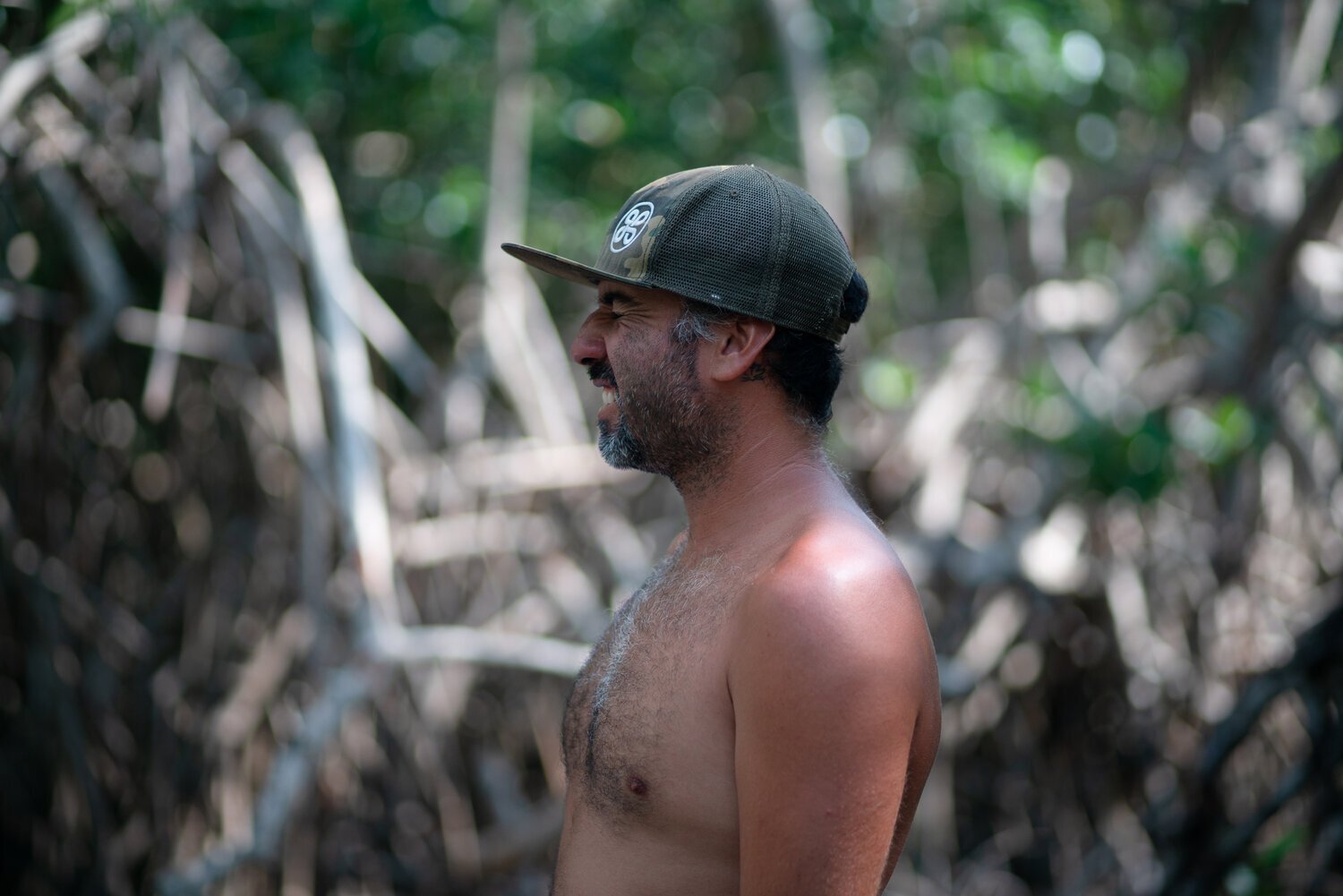 Homme barbu torse nu portant une casquette dans un environnement naturel avec des arbres.