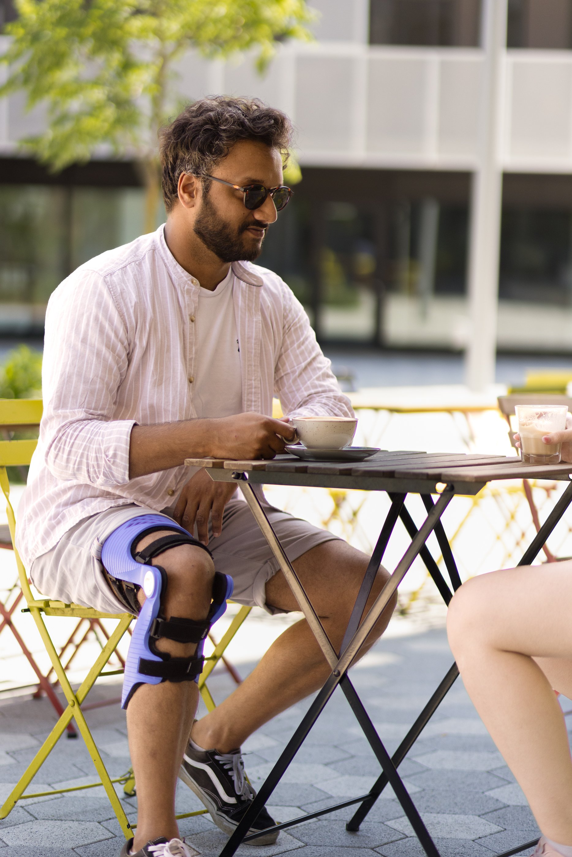 Un homme assis à une table extérieure, portant une attelle de genou et tenant une tasse. Il porte des lunettes de soleil et une chemise légère. Une autre personne assise en face est partiellement visible. La scène est en extérieur avec des arbres en 