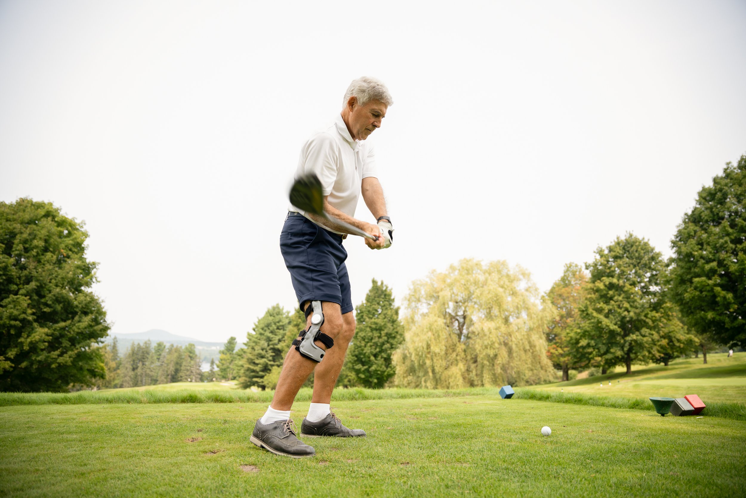 Un homme jouant au golf sur un parcours verdoyant, entouré d'arbres, tenant un club de golf et portant une orthèse au genou.